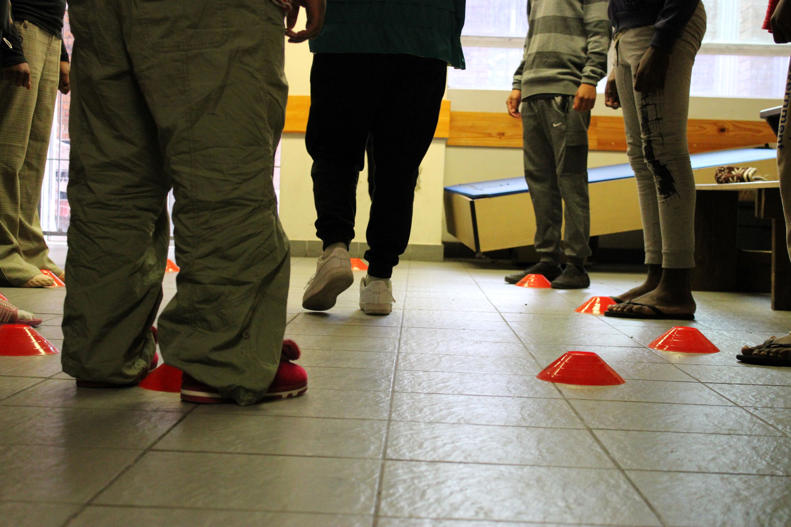 Children in a circle, shown from the waist down, participate in an activity involving orange cones