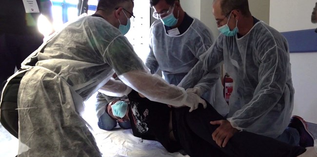 Τέσσερα άτομα με χειρουργικές ρόμπες και μάσκες εξασκούν μια τεχνική σε έναν εθελοντή ξαπλωμένο στο πλάι 