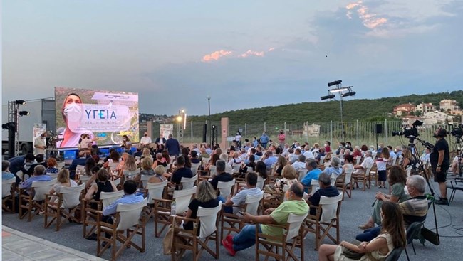 A seated audience in the open air faces a stage with seated panelists and a backdrop that reads "health" in Greek