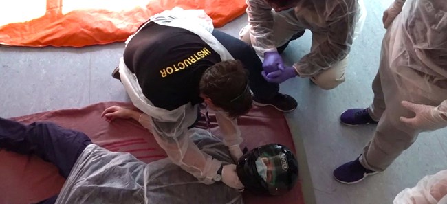 An instructor checks the pulse of a volunteer lying on the floor in a motorcycle helmet while trainees watch